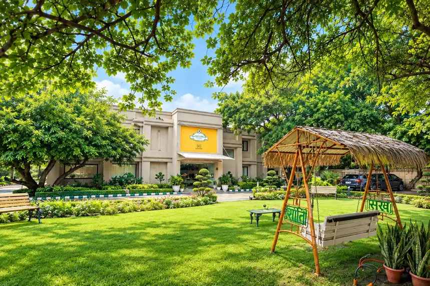 A lawn under spreading trees with a thatched swing seat, a garden bench and flower beds in front of a low cream building