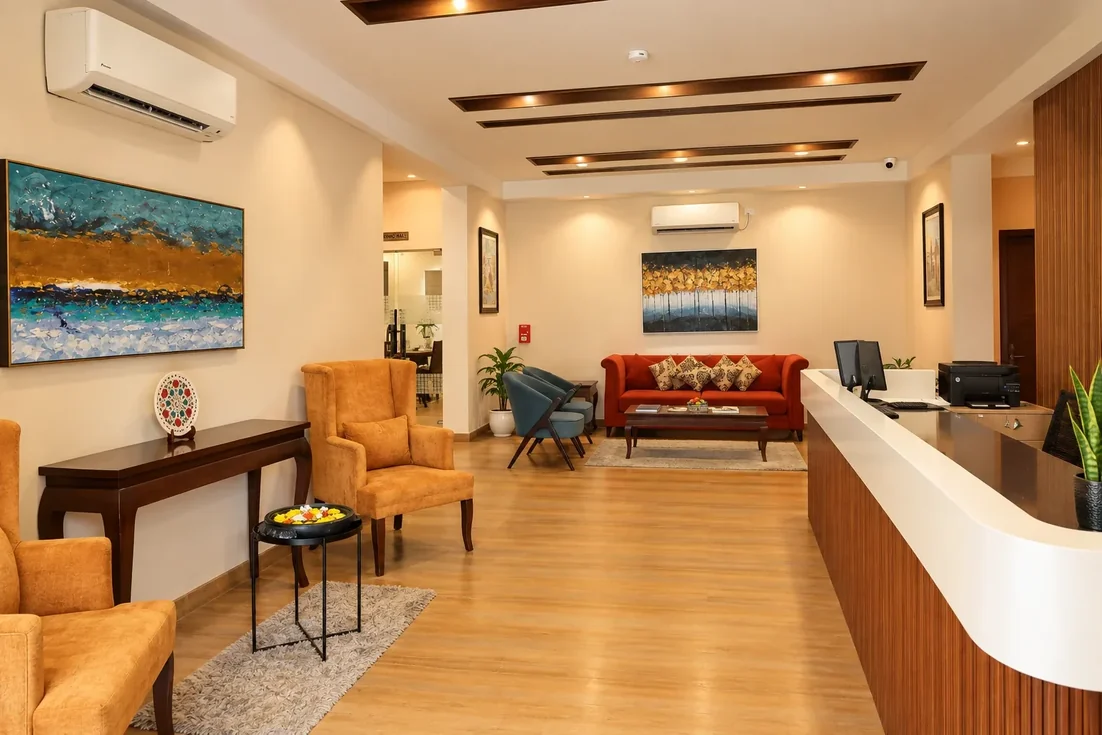 The reception at Vrindavan Staycation: a long curved desk in walnut and white, amber wing chairs, a red sofa under an abstract painting, and warm downlights in a coffered ceiling