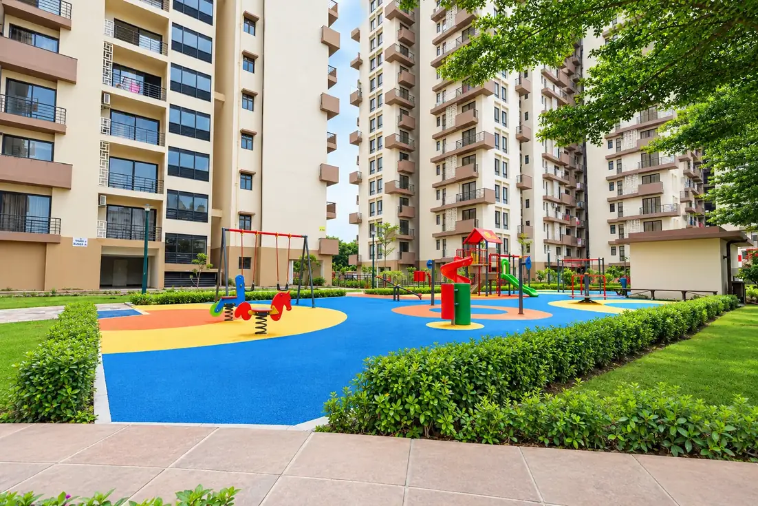 The children&rsquo;s play area between the apartment towers: swings, a slide and spring riders on a blue and yellow rubber surface, edged by hedges and lawn