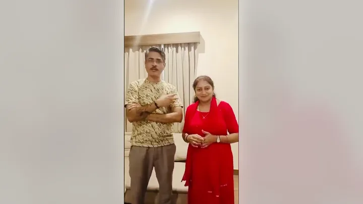 Video still: a couple standing in their room, speaking to the camera