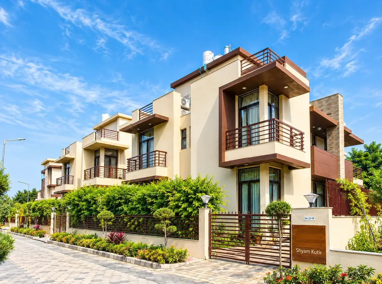 A villa at Krishna Bhumi: cream walls with rust-brown cantilevered balconies and a roof terrace, a slatted gate with the nameplate Shyam Kutir, clipped hedges and lamp posts along a paved lane, under a blue sky