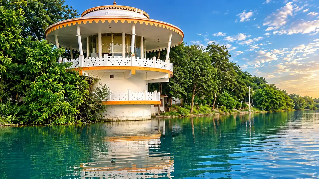 Devraha Baba&rsquo;s samadhi and ashram on the far bank of the Yamuna at Vrindavan: a round white pavilion with a saffron-trimmed canopy roof and lattice railings, set among trees at the water&rsquo;s edge and mirrored in the river