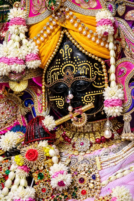The deity of Shri Radhavallabh, Vrindavan, in close-up: a black face painted with gold, a saffron turban strung with pearls, jasmine and rose garlands, jewelled ornaments and a golden flute at the lips