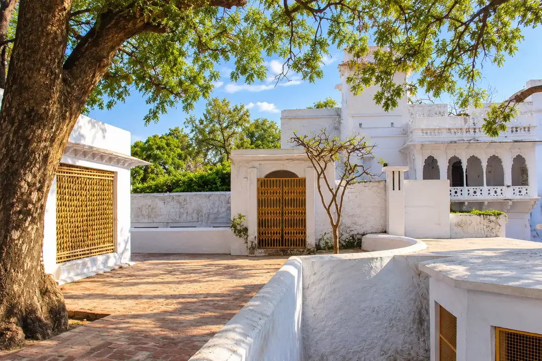 Tatiya Sthan, Vrindavan: a quiet brick-paved courtyard under a spreading tree, whitewashed walls with bamboo lattice screens and a saffron collapsible gate, and a white arched pavilion beyond